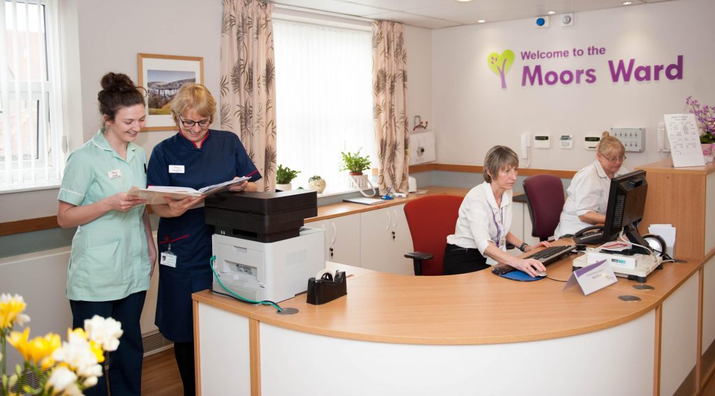 Moors Ward reception desk. Two receptionists are behind the desk and two nurses are standing at one end reading notes