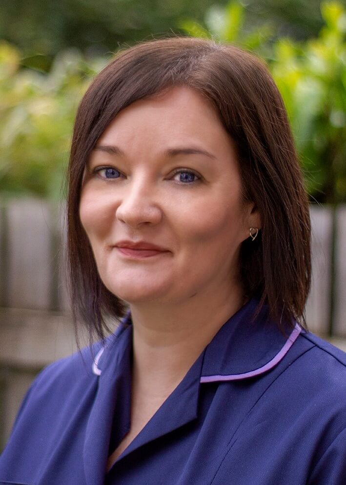 Head and shoulder portrait of a smiling nurse with dark brown hair wearing a dark blue uniform.