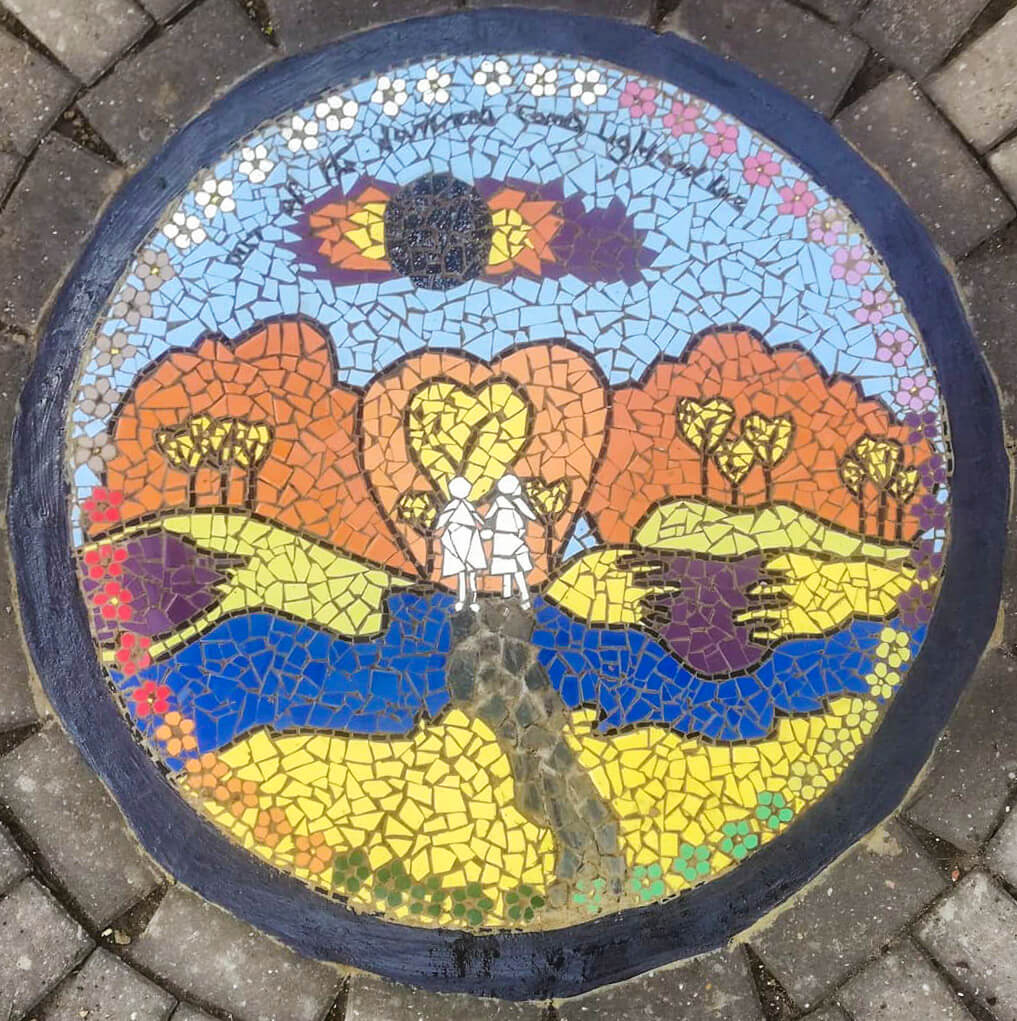A colourful circular mosaic depicting two people walking along a path towards trees. The words "Out of the darkness comes light and love" are written at the top of the mosaic.