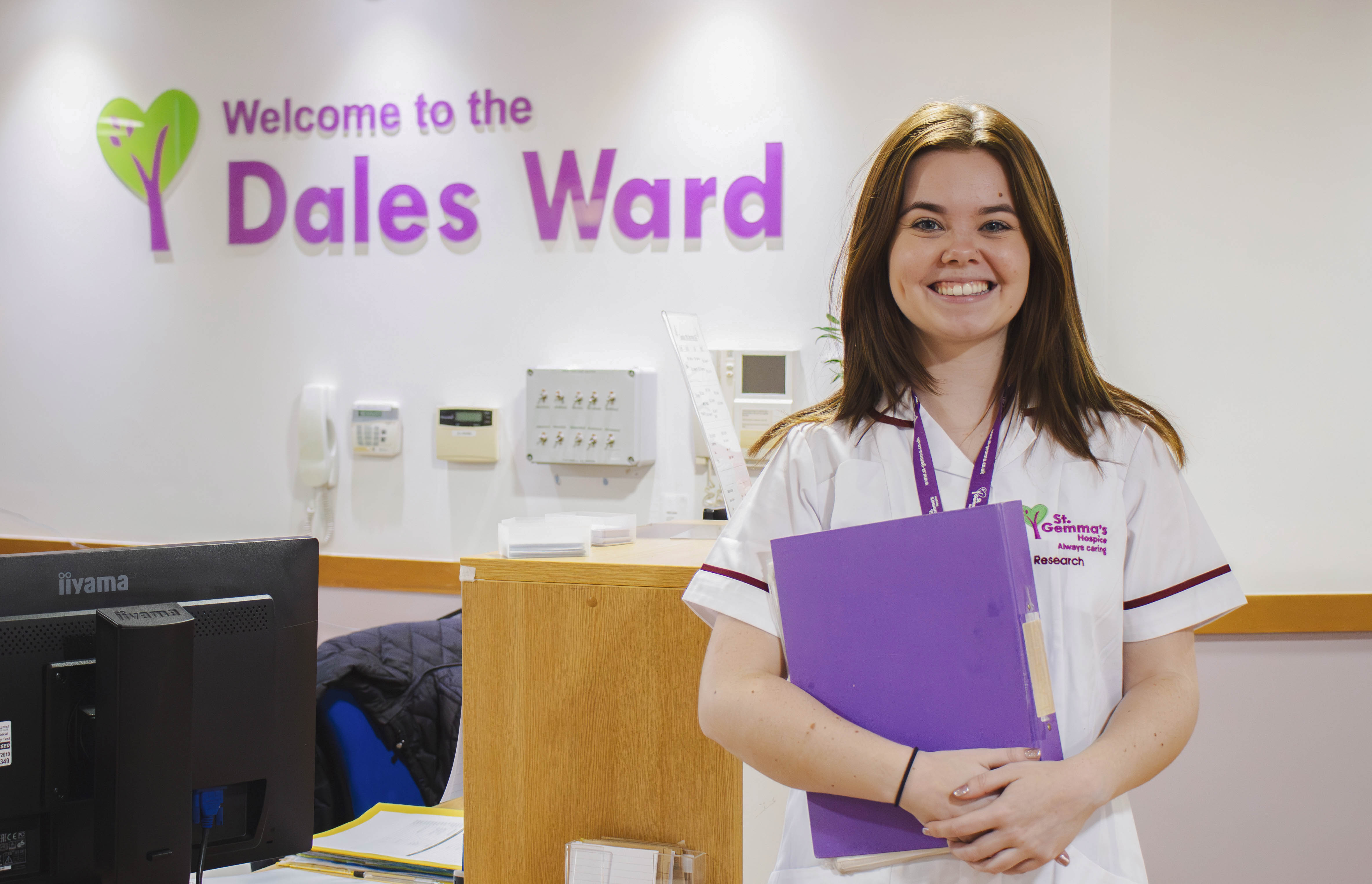 Woman wearing a white nurse's uniform is holding a purple folder and standing in front of a sign that reads Welcome to the Dales Ward